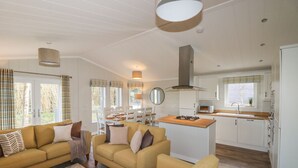 A modern living room with a sofa set, a dining area, and a kitchen with white cabinets and a wooden countertop. - 23 Meadow Retreat (Liskeard)