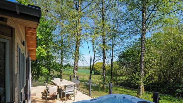 A hot tub on a wooden deck with a table and chairs, surrounded by trees and a view of the countryside. - 23 Meadow Retreat (Liskeard)