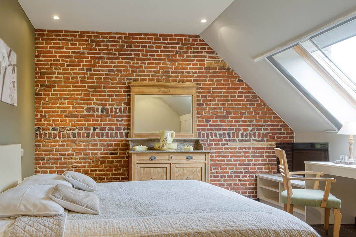 Un dormitorio con techo inclinado, pared de ladrillo, cama con almohadas, cómoda de madera, espejo y escritorio con silla.
