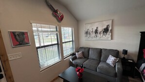 Un salon avec un canapé gris, une table basse ornée d’un vase de fleurs, une guitare accrochée au mur et un tableau représentant des chevaux.