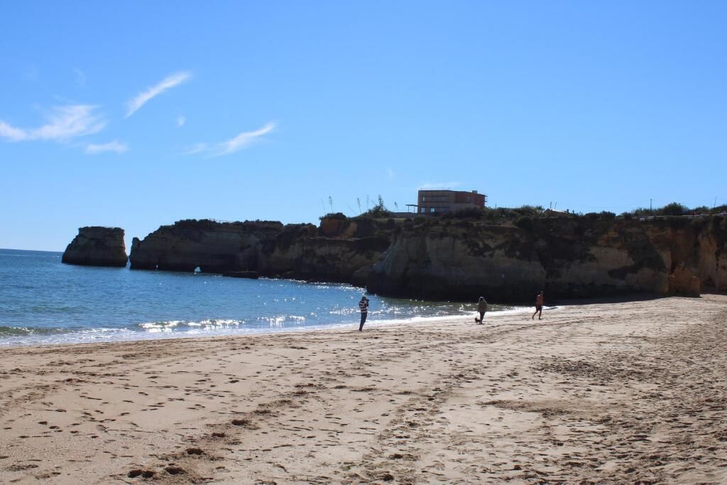 Una playa con un edificio en un acantilado, cielo azul despejado y huellas en la arena.
