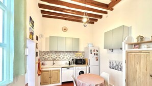 A compact kitchen with a dining area, wooden ceiling beams, a washing machine, and a refrigerator. - Au Croco fleuri, 50m des arènes, terrasse (Nîmes)