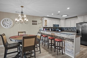 Una cocina con armarios blancos, un refrigerador de acero inoxidable, una mesa de comedor con sillas y una barra con taburetes.