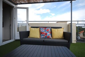 Un balcón exterior con un sofá de mimbre, dos cojines amarillos y una mesa.