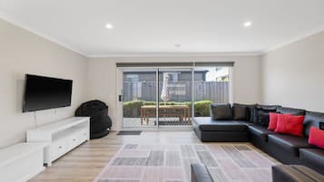 A modern living room with a flat-screen TV, a black bean bag, a black leather sofa with red cushions, a white console, and a patterned rug.