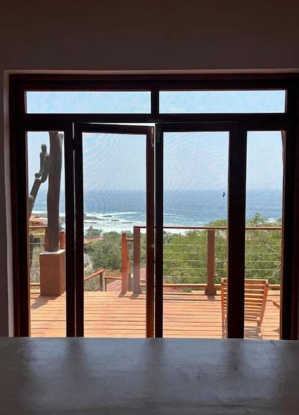 A view through a large window of a wooden deck overlooking a coastal landscape with a statue and a clear sky.