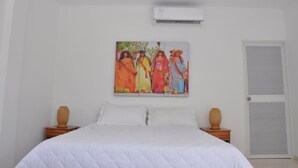 A bedroom with a bed, a painting on the wall, a door, and an air conditioning unit.