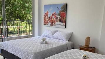 A hotel room with two beds, a balcony with a view of greenery, and a painting on the wall.
