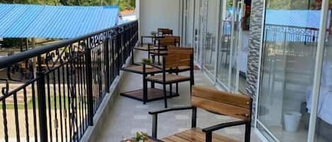 A balcony with wooden tables and chairs, a pool, and a view of palm trees and a blue-roofed building.