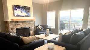 A living room with a stone fireplace, a television mounted above it, a sofa, a coffee table, and a view through large windows. - Unwind and relax at this luxurious mountain retreat. Beautiful Views! (Sparta)