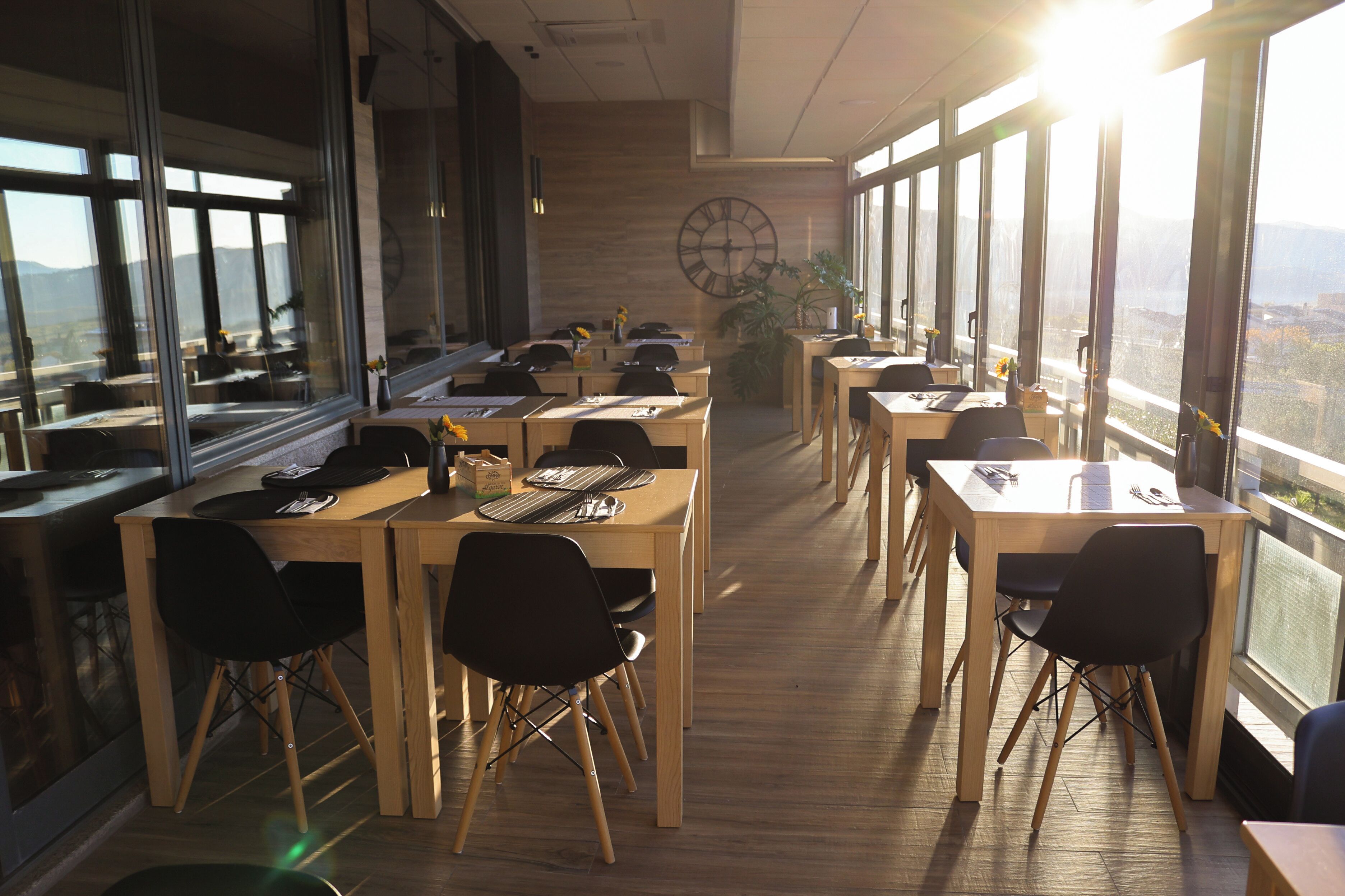 Een moderne eetruimte met houten tafels en zwarte stoelen, grote ramen met uitzicht op de stad, en een grote wandklok.