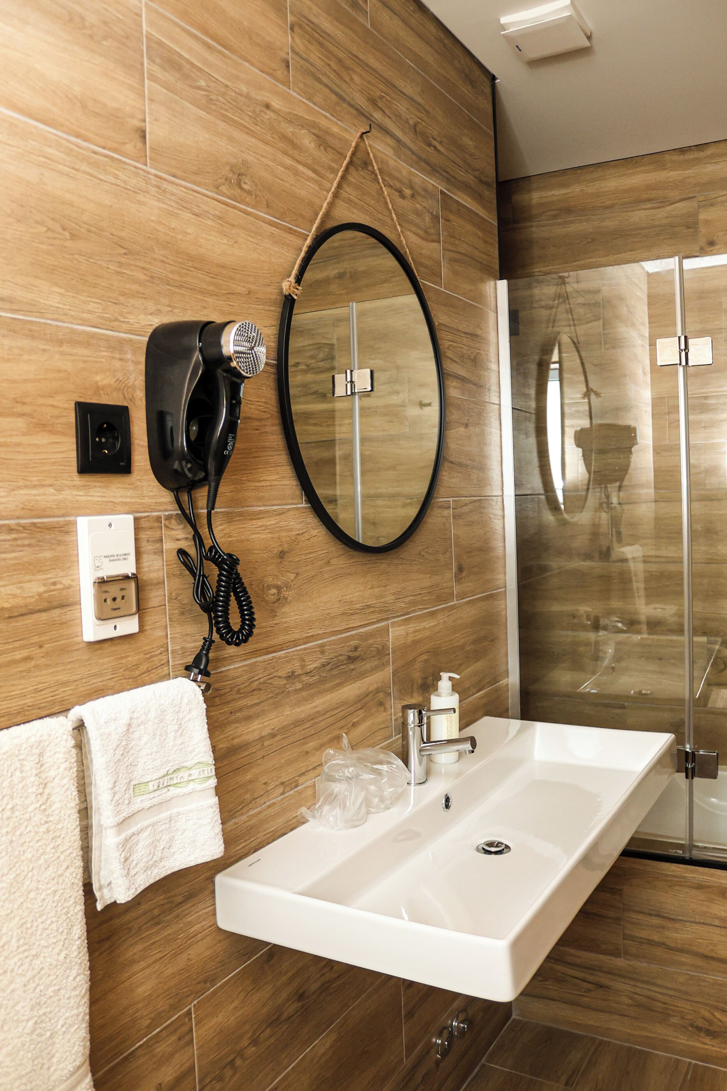 A bathroom with a wooden wall, a white sink, a hairdryer, a mirror, and a glass shower enclosure.
