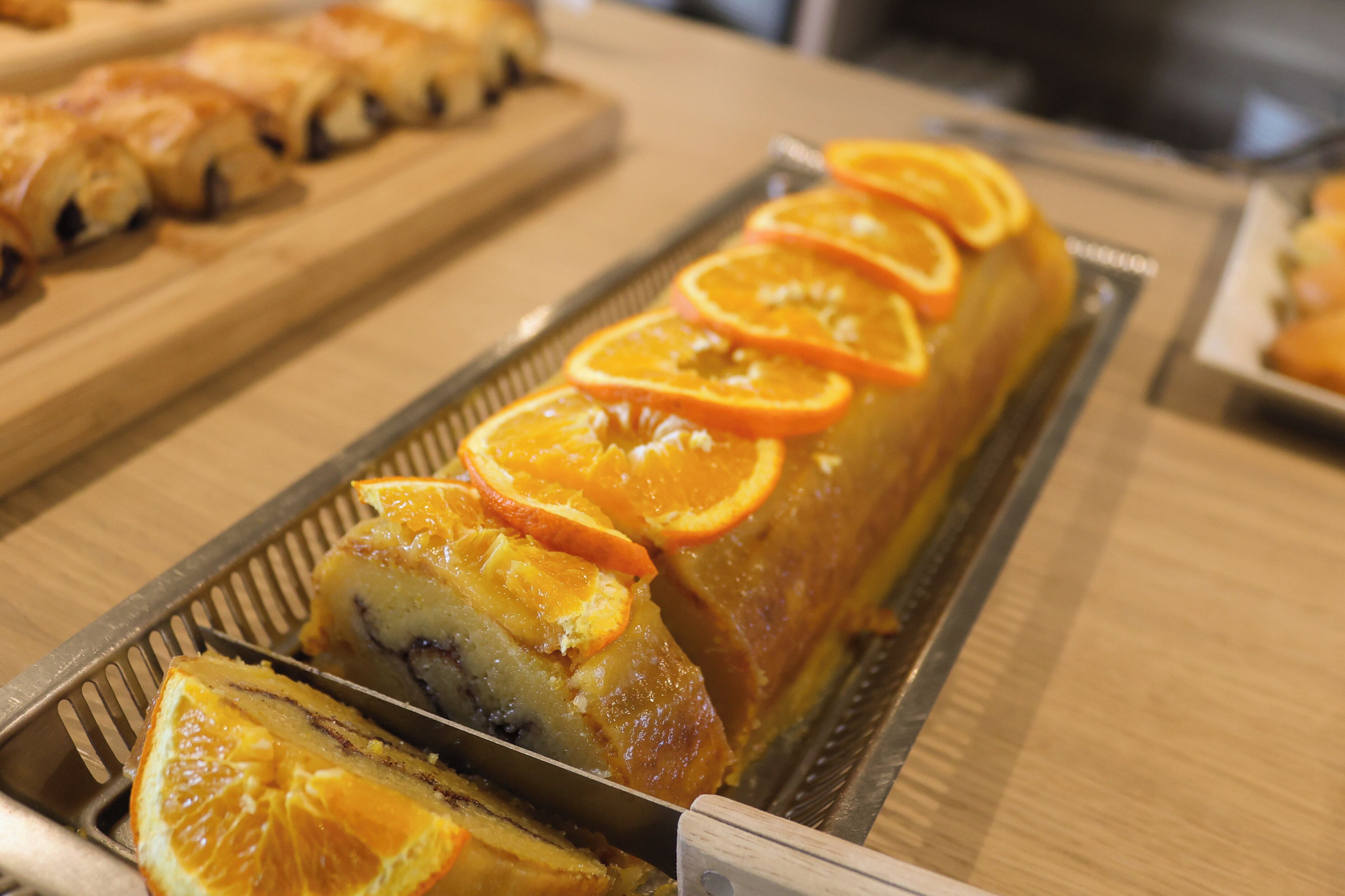 A long, rectangular pastry with orange slices on top, placed on a metal tray.