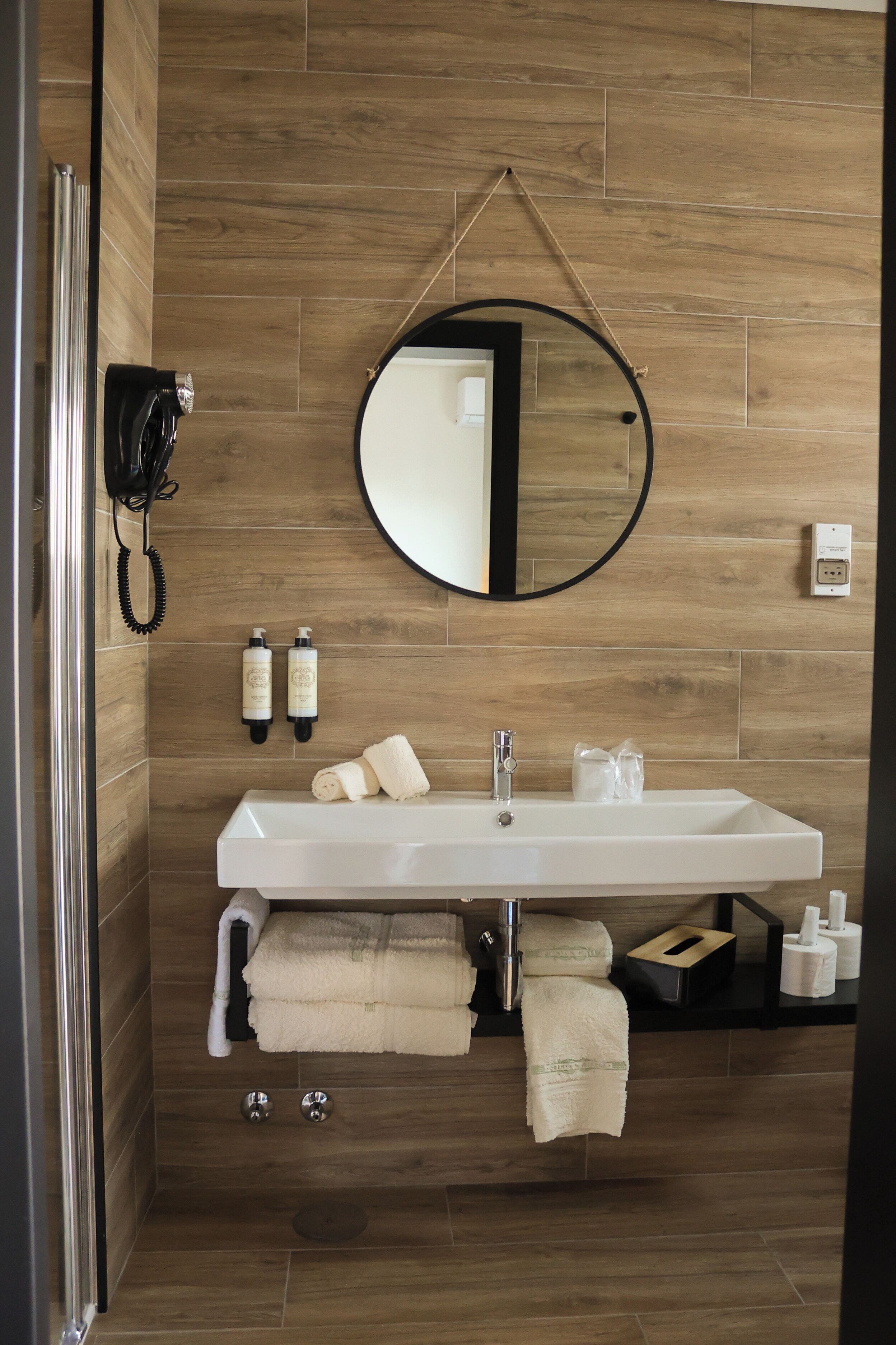 A modern bathroom with a wooden wall, a large mirror, a white sink, and two bottles of shampoo.