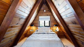 A cozy attic bedroom with a sloped ceiling, wooden walls, a bed with a checkered duvet, two bedside tables with lamps, and a window providing natural light.