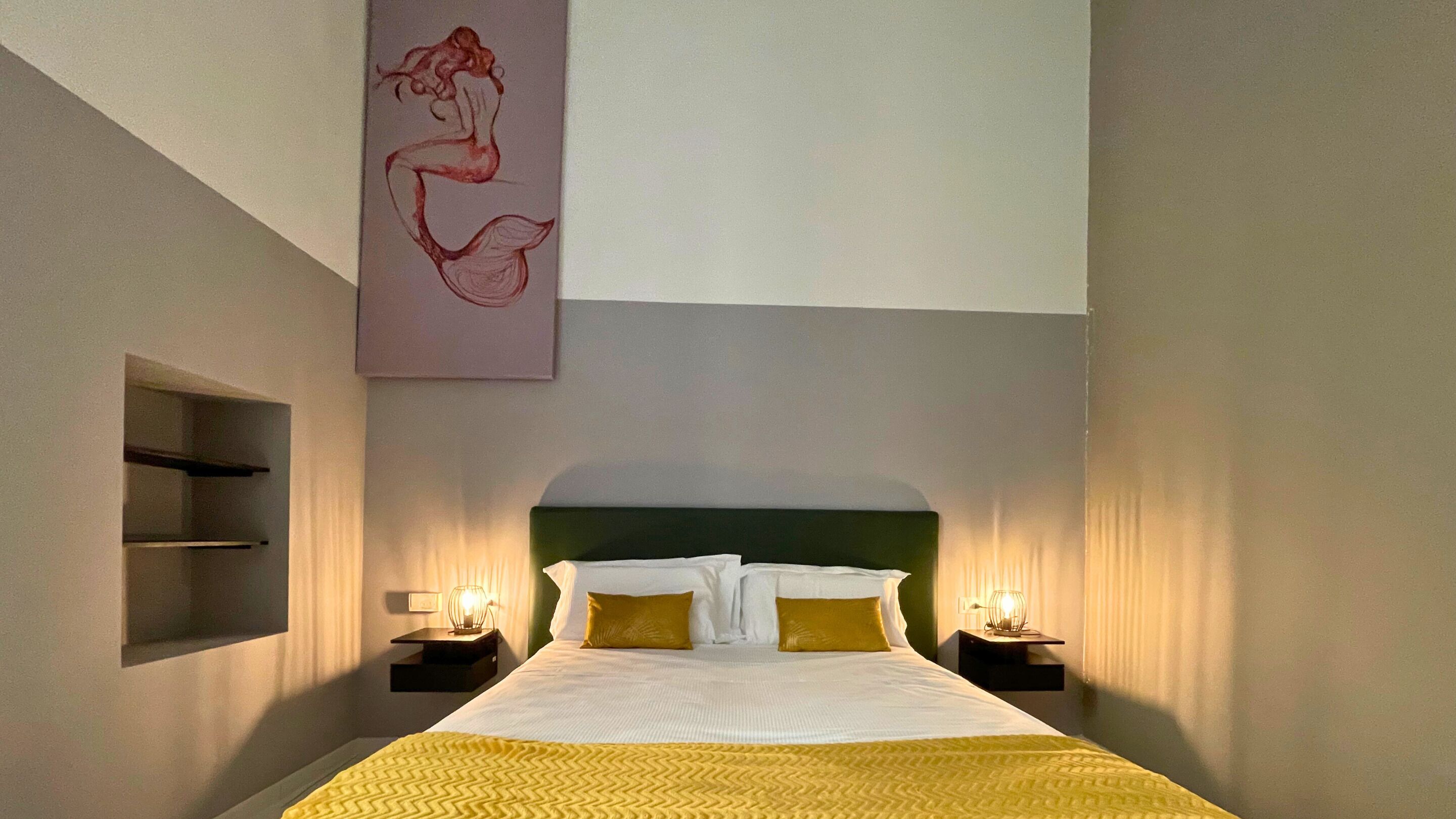 Habitación de hotel con una cama grande, dos mesitas de noche con lámparas, un estante en la pared y un cuadro abstracto de gran tamaño.