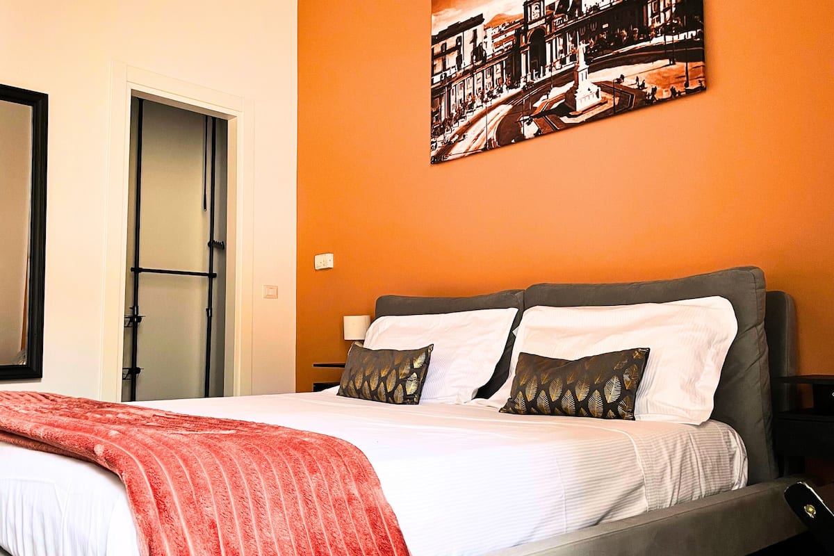 A hotel room with a bed, a door, and a wall with a cityscape painting.