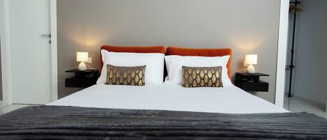 Un lit bien fait, avec une literie blanche et orange, deux coussins décoratifs et un plaid noir.