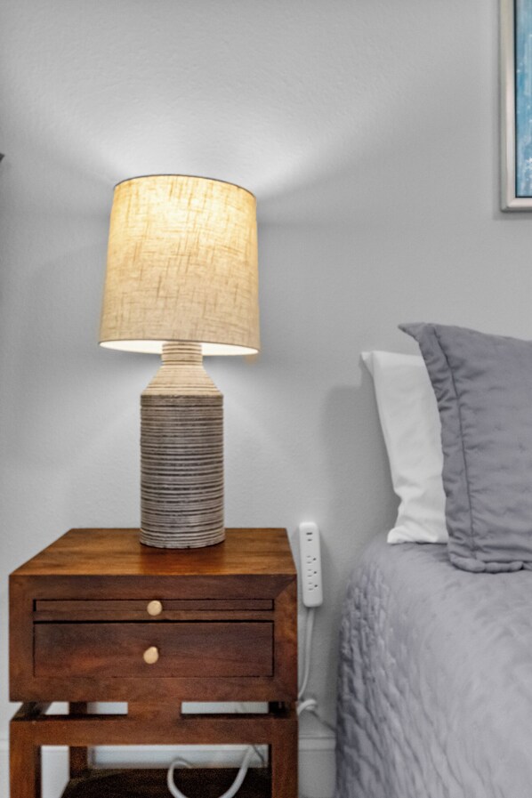 Una mesita de noche de madera con una lámpara y un enchufe, una lámpara de noche blanca y una cama con una manta gris y almohada blanca.