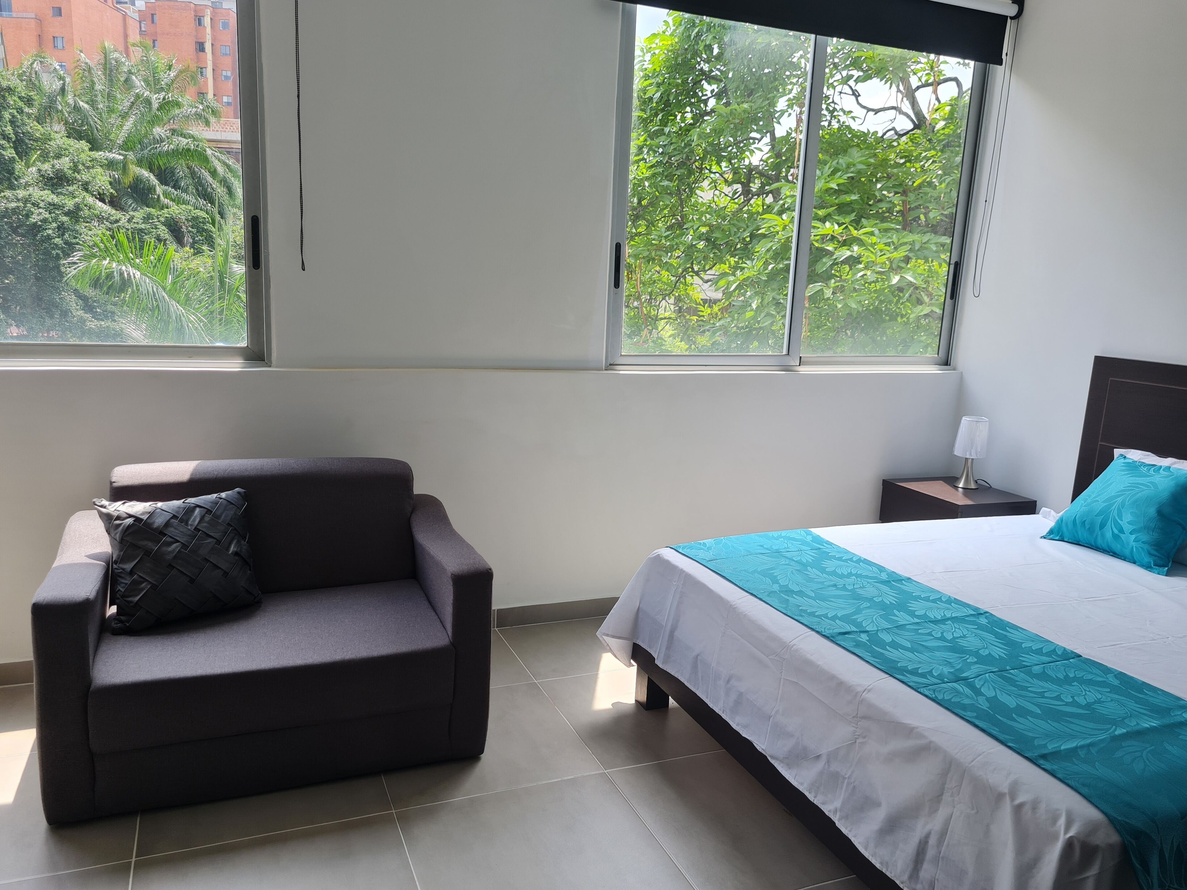 A hotel room with a bed, a sofa, a nightstand, and a window with a view of greenery and buildings.