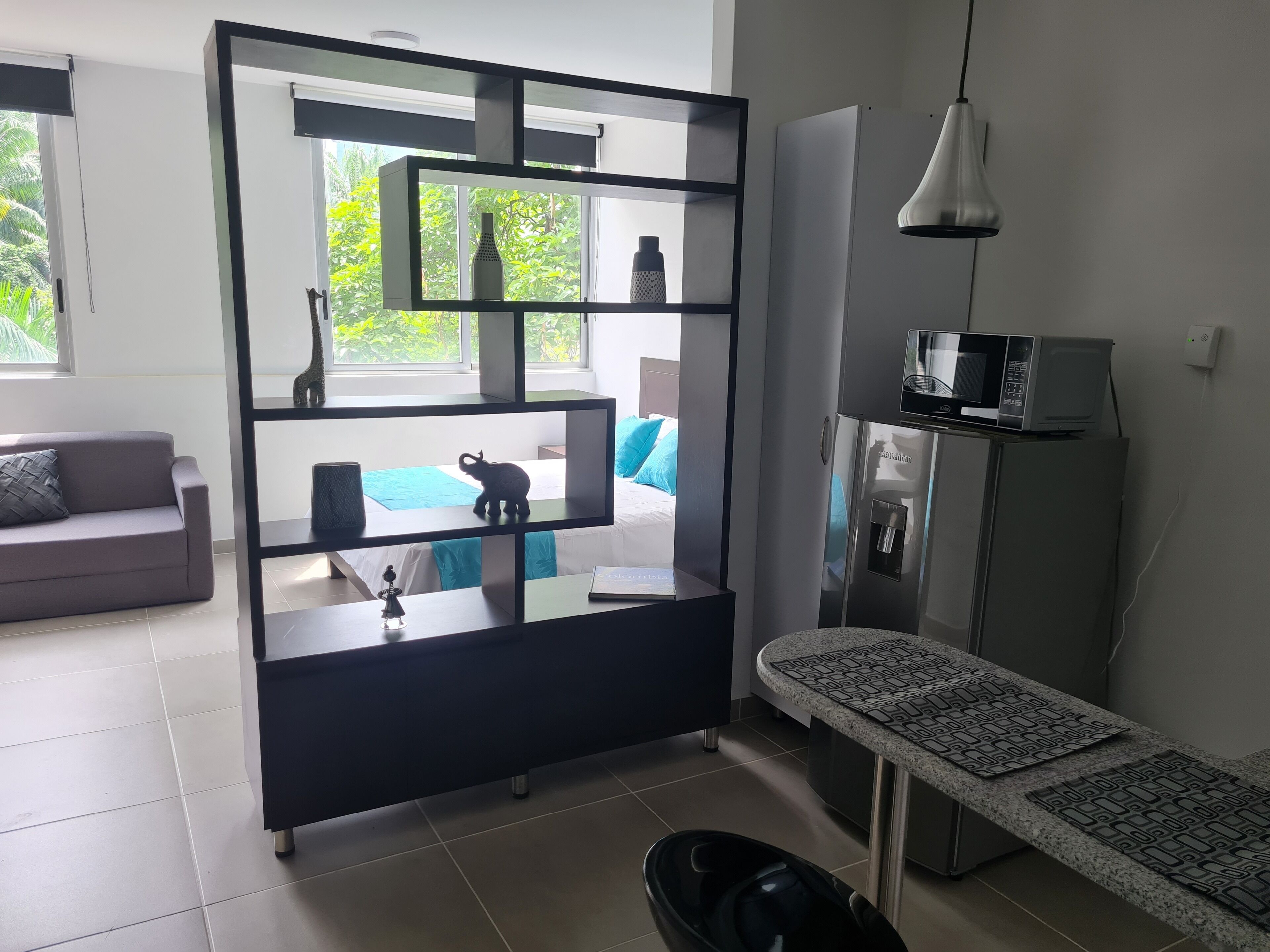 A modern living room with a sofa, a coffee table, a glass partition with shelves, a bench, a microwave, and a water dispenser.