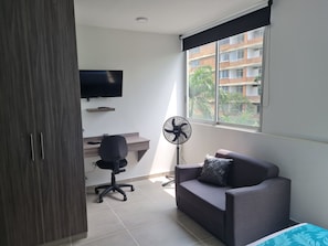 Een moderne kamer met een grijze bank, een zwarte bureaustoel, een aan de muur bevestigde televisie en een raam met uitzicht op gebouwen.