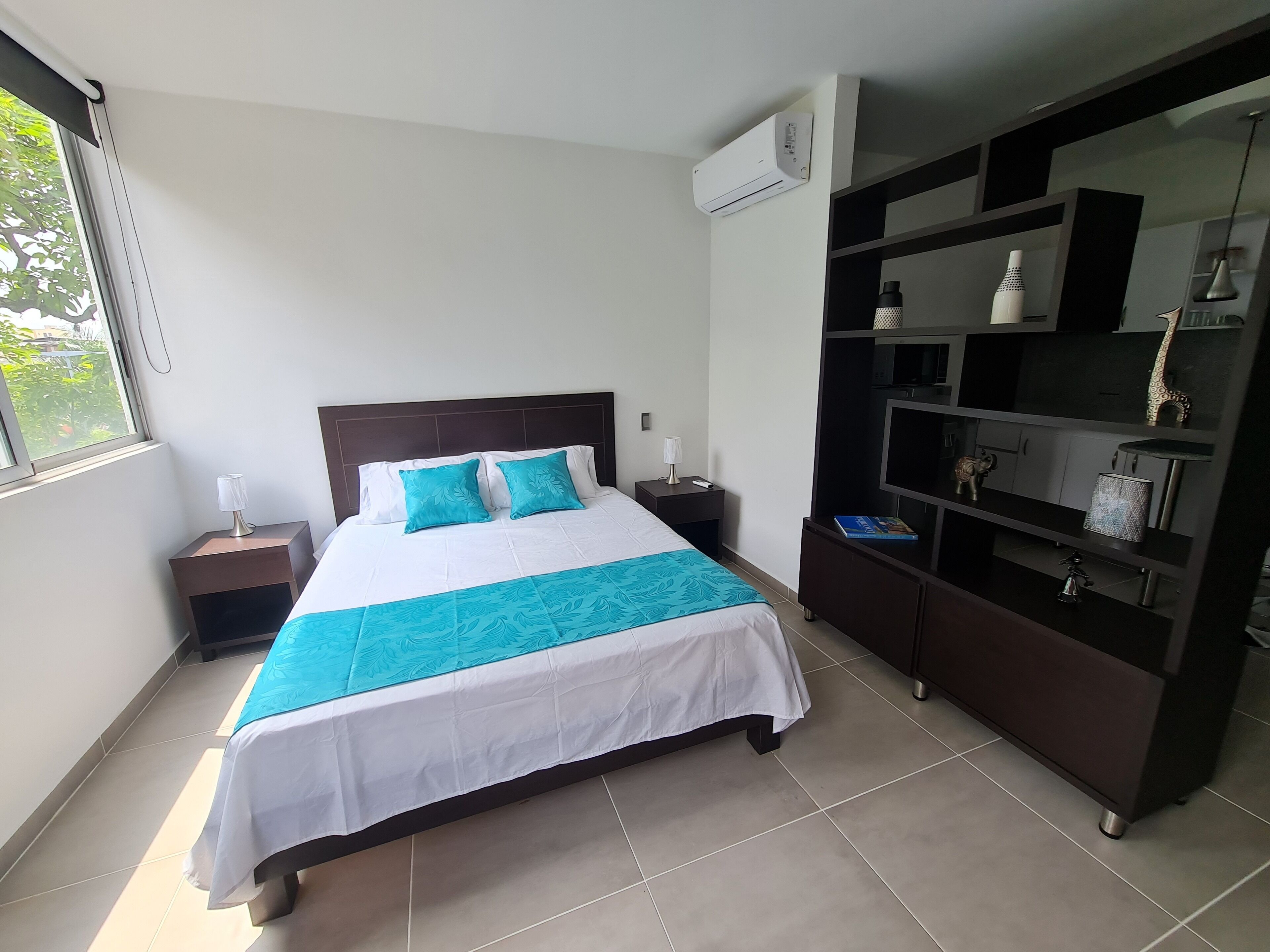 A neatly arranged hotel room with a bed, a nightstand, a lamp, a TV on a cabinet, and a window with a view of greenery.