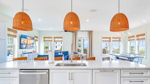 A modern kitchen with white cabinets, a stainless steel sink, and a large island. There are wicker pendant lights, a flat-screen TV, and a dining area with a table and chairs. - The Sand Pearl-New/N Cape/Steps to Beach/5 BR (Port St. Joe)