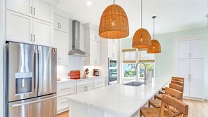 A modern kitchen with white cabinetry, a stainless steel refrigerator, a large island with bar stools, and wicker pendant lighting. - The Sand Pearl-New/N Cape/Steps to Beach/5 BR (Port St. Joe)