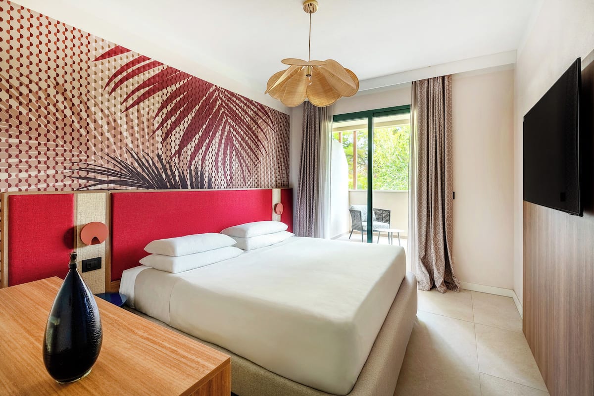 Ein Hotelzimmer mit einem großen Bett, einem Nachttisch aus Holz, einer dekorativen Wandverkleidung, einer Deckenleuchte, einem Fernseher und einem Balkon mit Vorhängen.