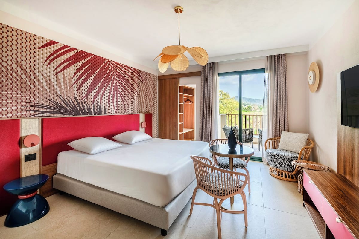 Ein Hotelzimmer mit Bett, einem kleinen Tisch, Korbstühlen, einem Fernseher und einem Balkon mit Blick.