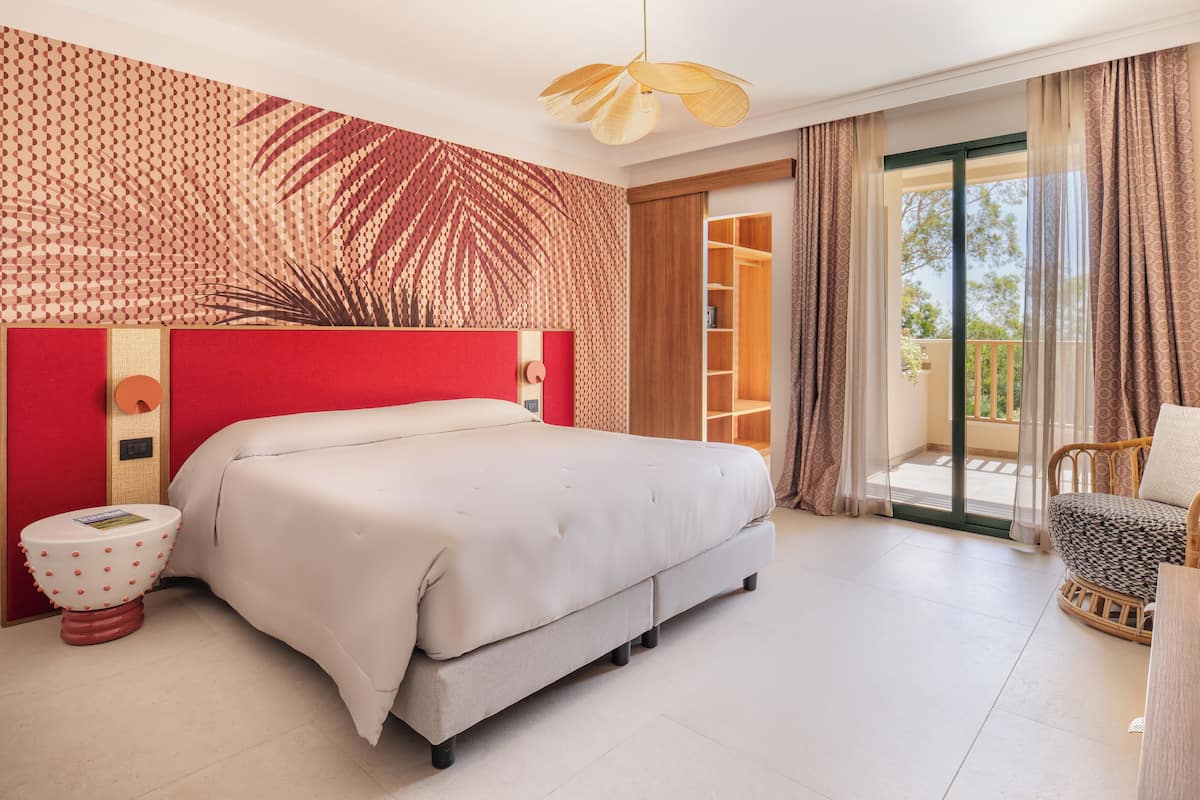 Ein Schlafzimmer mit einem großen Bett, einer roten Akzentwand, einem hölzernen Kopfteil, einem kleinen runden Tisch und einem Balkon mit einem Korbstuhl.