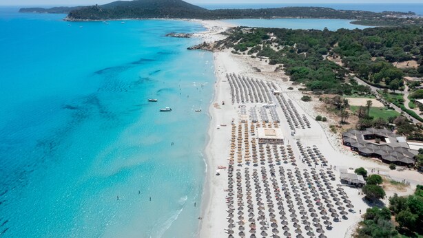 An aerial view of a beach with rows of umbrellas and beach chairs, a sandy shoreline, and a distant island. - VOI Tanka Selected (Villasimius)