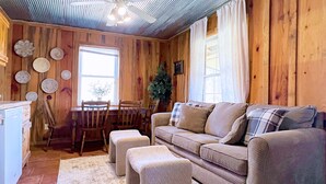 Ein gemütliches Wohnzimmer mit einem beigen Sofa, passenden Hocker, einem Holzesstisch mit Stühlen und einem großen Fenster mit Vorhängen.