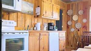 Eine Küche mit Holzwandverkleidung, einer Mikrowelle, einem Herd und einem Geschirrspüler. An der Wand befinden sich gewebte Untersetzer und es steht ein Stuhl in der Ecke.