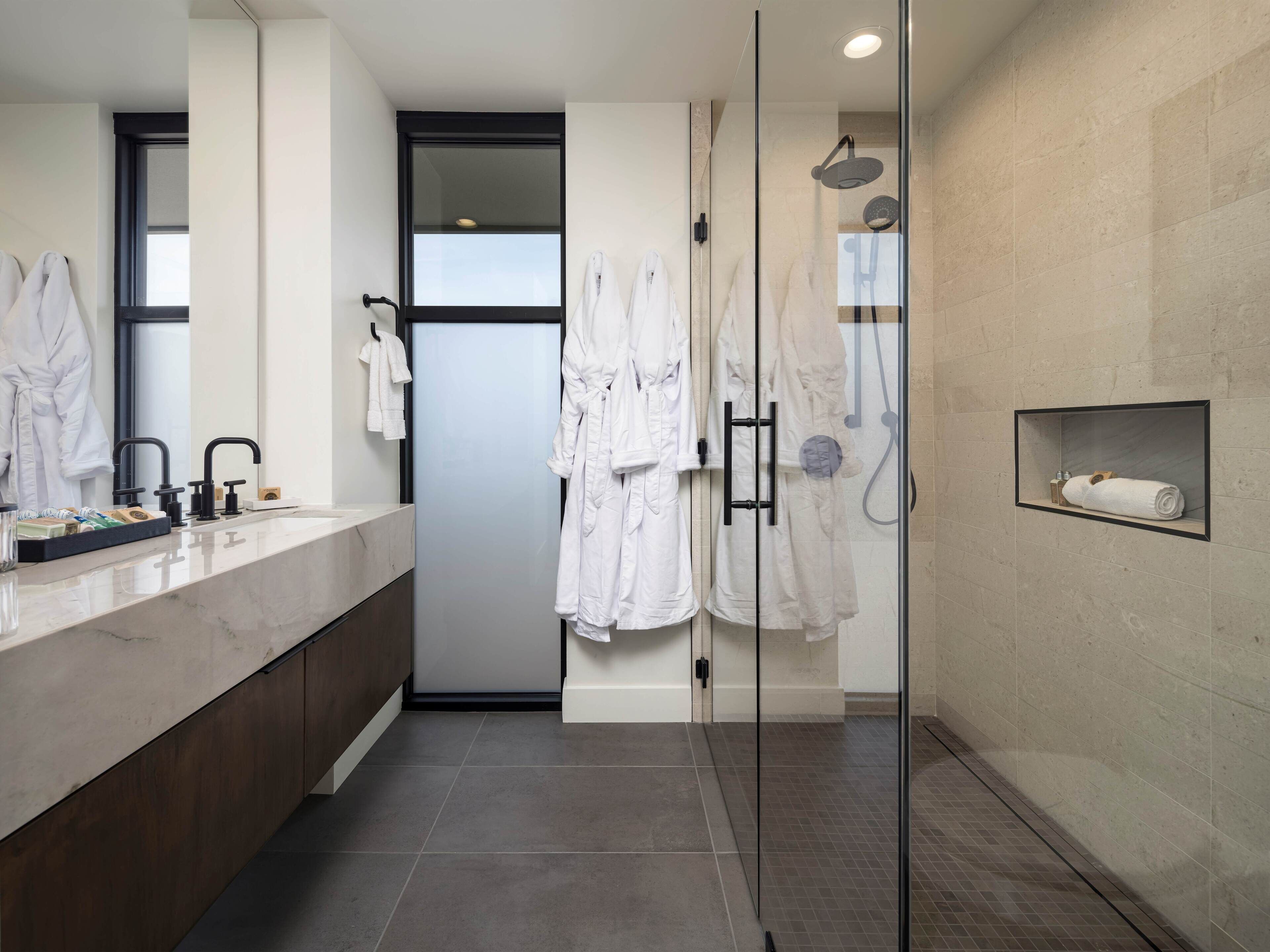 Un baño moderno con ducha de vidrio, tocador de mármol y varias batas blancas colgadas en ganchos.