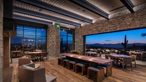 Un vaste espace extérieur aménagé pour les repas, avec des tables et des bancs en bois, des piliers en pierre et de grandes fenêtres offrant une vue sur les montagnes.