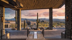 Un espace extérieur abrité, doté de piliers en pierre, d’un banc en béton et offrant une vue sur un paysage désertique, avec des cactus et des montagnes en arrière-plan.