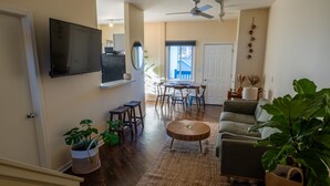 Un salon comprenant un canapé, une table basse et un coin repas avec des chaises. Un téléviseur est fixé au mur, il y a un ventilateur de plafond et une grande fenêtre offrant une belle vue.