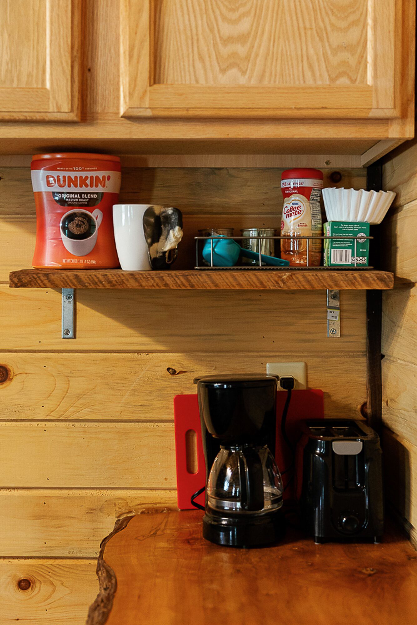 Een keuken met houten kastjes, een koffiezetapparaat, een koffiekan, een mok, een blik Dunkin' koffie, een potje room en een doos papieren bekertjes.