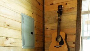 Een houten cabin met een houten wand, een gitaar die aan de wand hangt, een grijze bank met kussens en een raam.