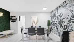 A modern dining area with a round table, black chairs, and a glass top. There is a green wall with a decorative light fixture, a white bench, and a wall with graffiti-style writing.