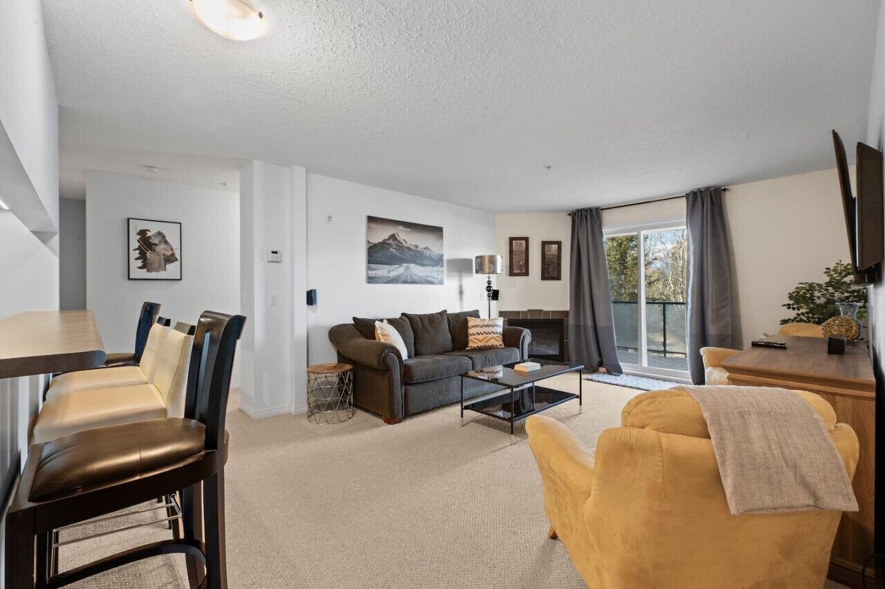 A modern living room with a sofa, coffee table, and chairs. There is a fireplace and a large window with curtains.
