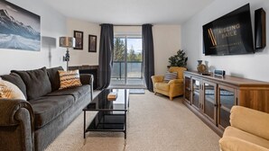 Una sala de estar moderna con un sofá gris, un sillón amarillo, una mesa de centro de vidrio y un televisor montado en la pared.
