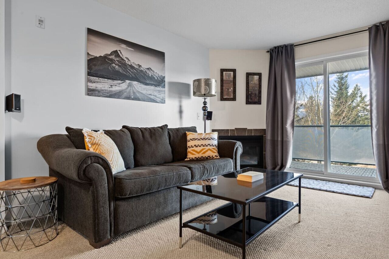 A modern living room with a grey sofa, a coffee table, and a fireplace. There is a large window with curtains, and a mountain landscape painting on the wall.