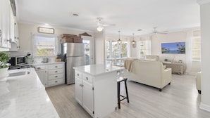 A spacious kitchen with a central island, a dining area with a sofa, and a TV mounted on the wall. - Beach House - Private Dock + Walk to the Beach (Holden Beach)