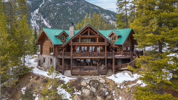 A log cabin with a green roof, multiple balconies, and a stone foundation, surrounded by trees and mountains.