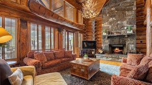 A cozy log cabin interior with a stone fireplace, wooden furniture, and large windows.