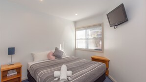 Ein Schlafzimmer mit einem Bett, einem an der Wand befestigten Fernseher, einem Fenster mit Jalousien und einem Nachttisch mit einer Lampe.