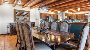 Une salle à manger avec une table et des chaises en bois, un lustre et, au fond, une cuisine.